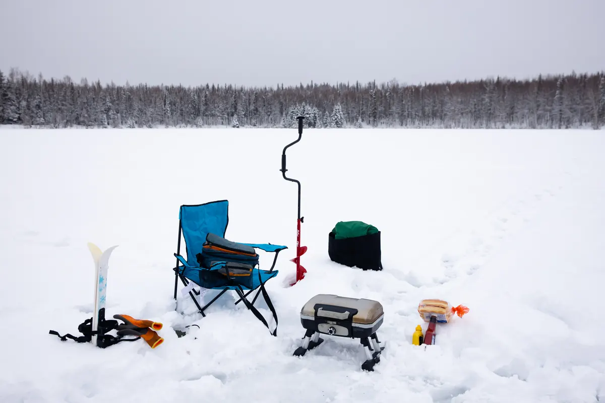 Ice fishing gear, a folding chair and an auger set up on a frozen lake ringed by spruce