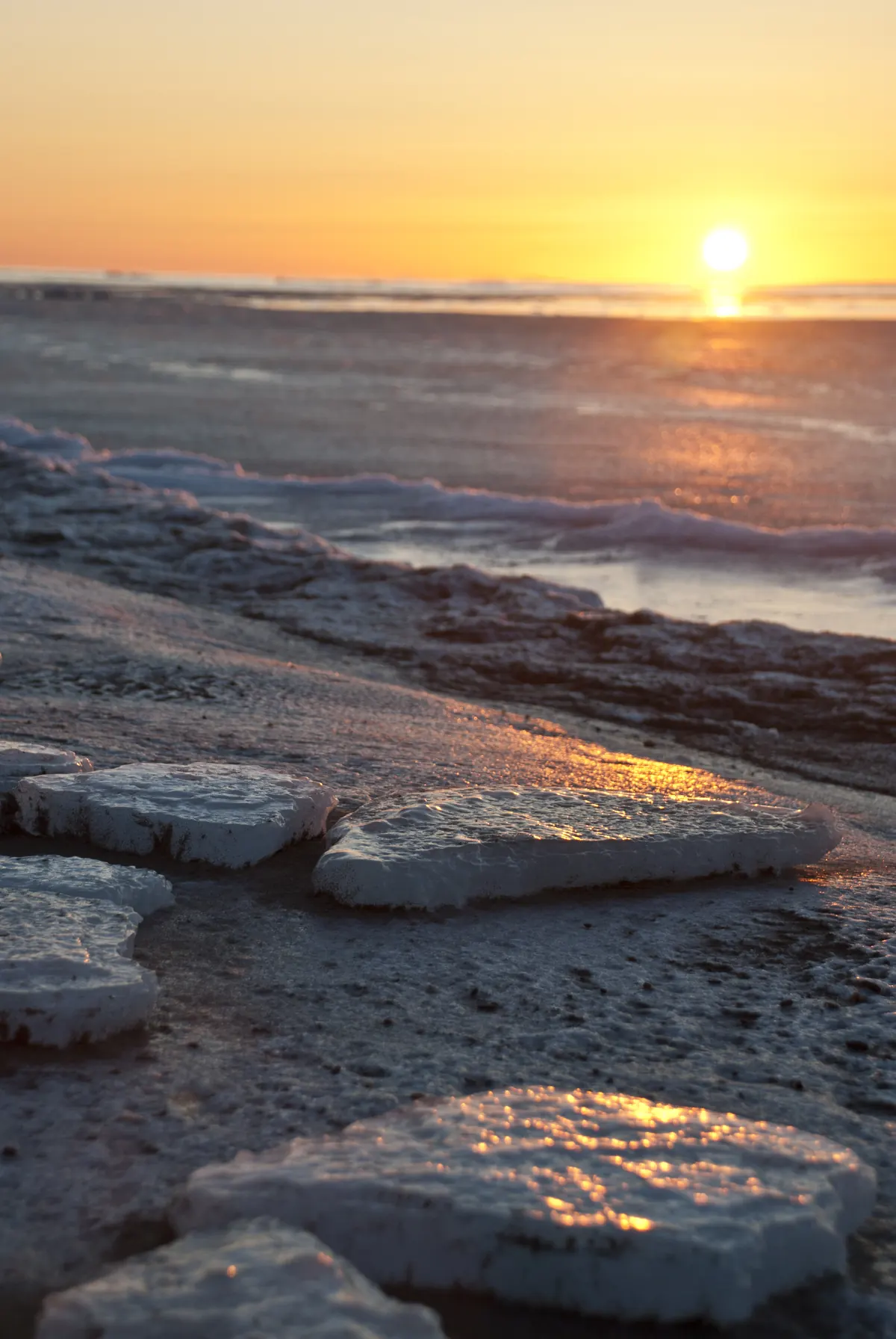 Flat plates of new sea ice washed up on a dark beach with the sun low over the Bering Sea at sunrise