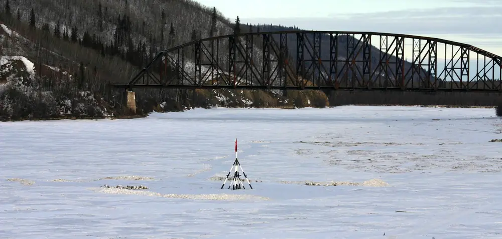 A red-flagged wooden tripod standing on the frozen Tanana River below a steel railroad bridge