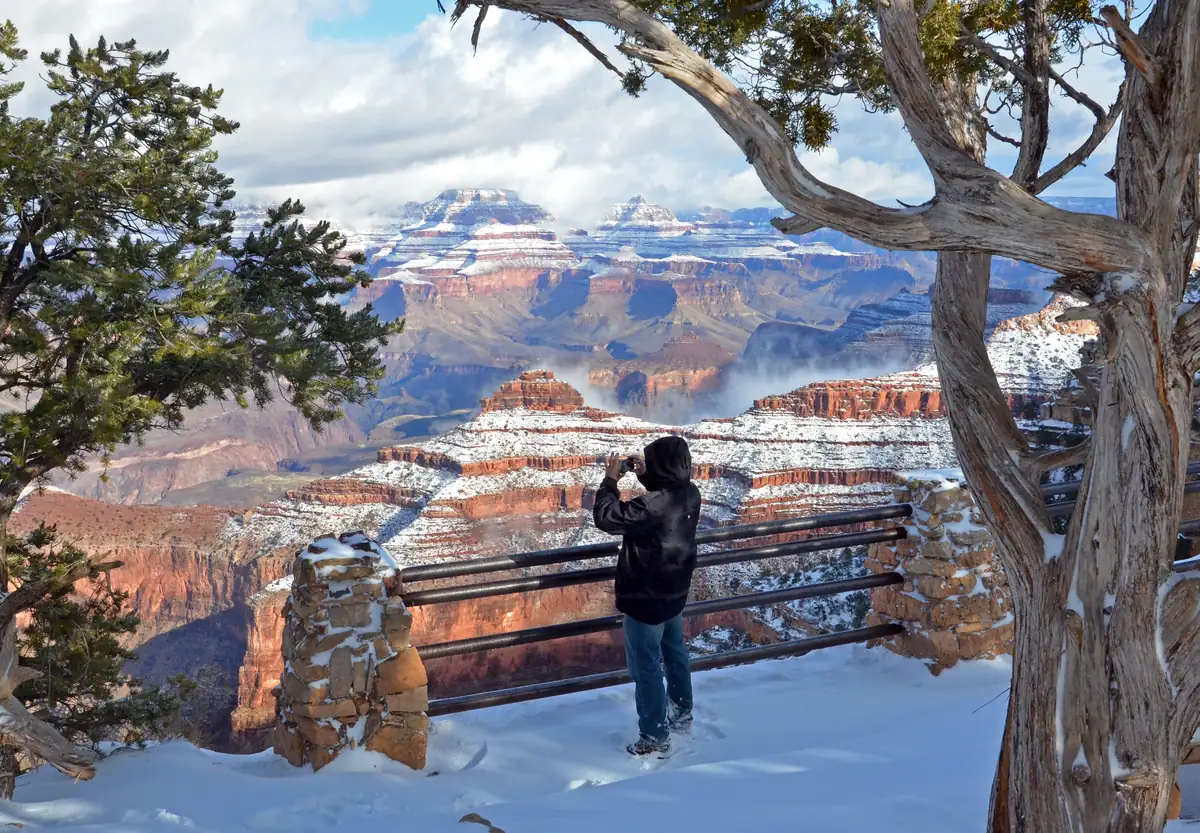 A visitor in a dark coat photographing the snow-dusted buttes of the Grand Canyon from a snowy overlook framed by juniper and pinyon trees.
