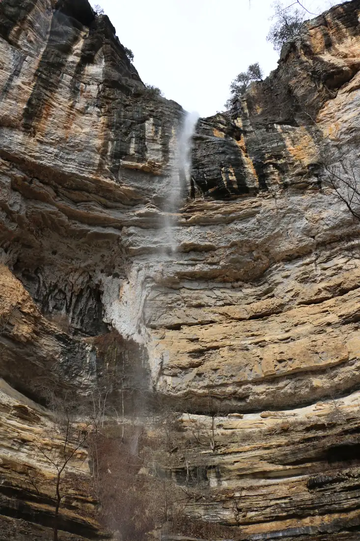 A tall narrow waterfall blowing sideways in the wind as it drops off a sandstone bluff