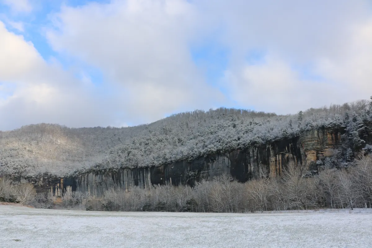 A long gray bluff and wooded ridge above a snow-covered field, with frosted bare trees under a partly cloudy sky.