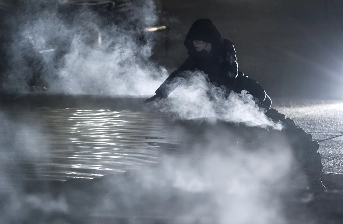 Thick white steam rising from a pool of thermal water at night, with a person in a dark hooded coat reaching toward the water