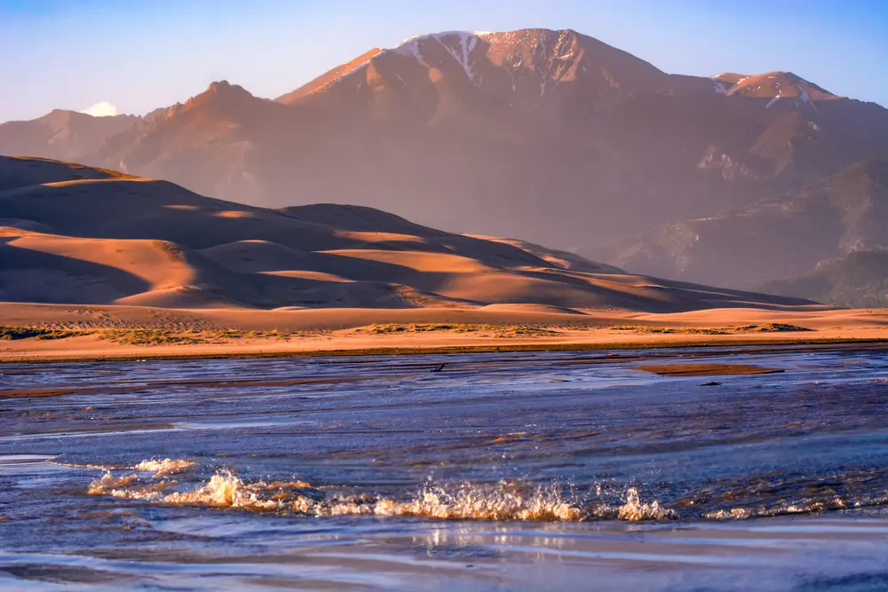 Waves of Medano Creek rippling across sand with the dunes and Sangre de Cristo peaks behind