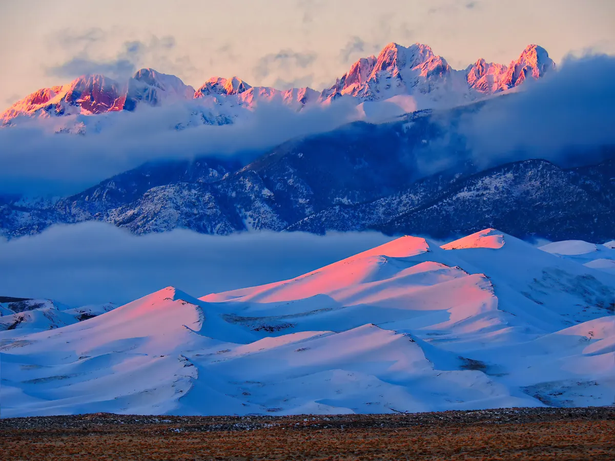 Snow-covered sand dunes glowing pink at sunrise below cloud-wrapped, snowy mountain peaks.
