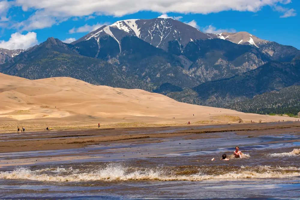 A wave of brown water rolling across the sand of Medano Creek in front of the Great Sand Dunes and snow-streaked Sangre de Cristo peaks, with a few people wading
