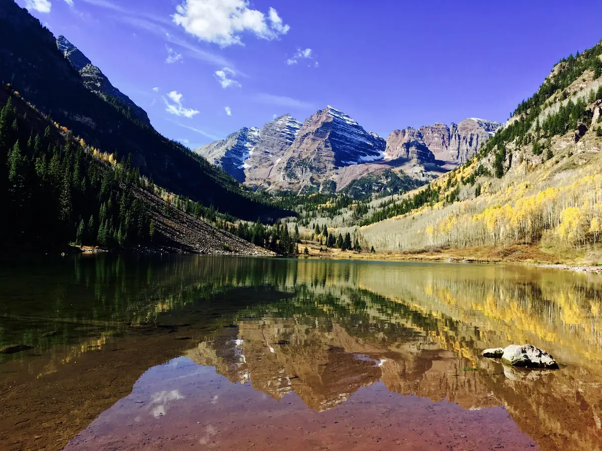 Yellow aspens and dark conifers below the Maroon Bells peaks, reflected in a still lake under a deep blue sky.