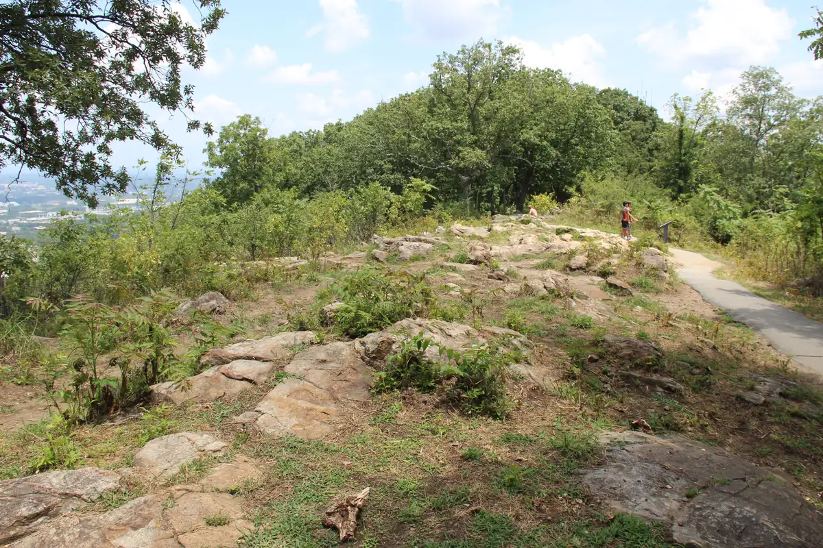 Rocky, open summit of Kennesaw Mountain with a paved path, low oaks and a distant view of the suburbs to the left