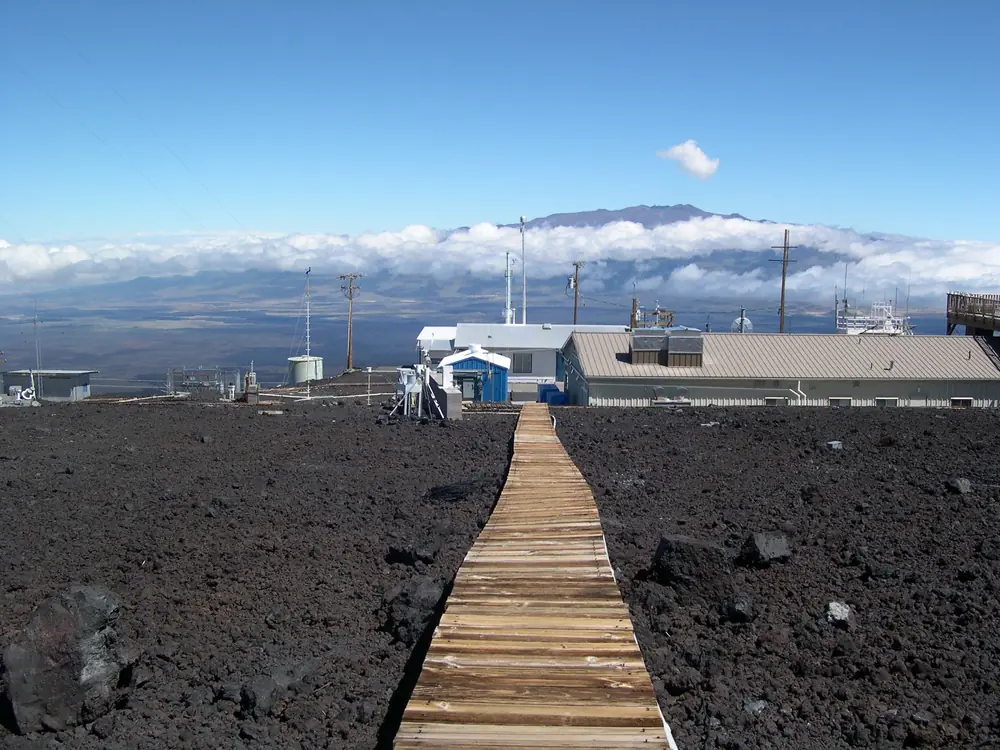Low buildings and instrument towers of an observatory stand on black lava rock beneath clouds, with a long wooden boardwalk leading to them
