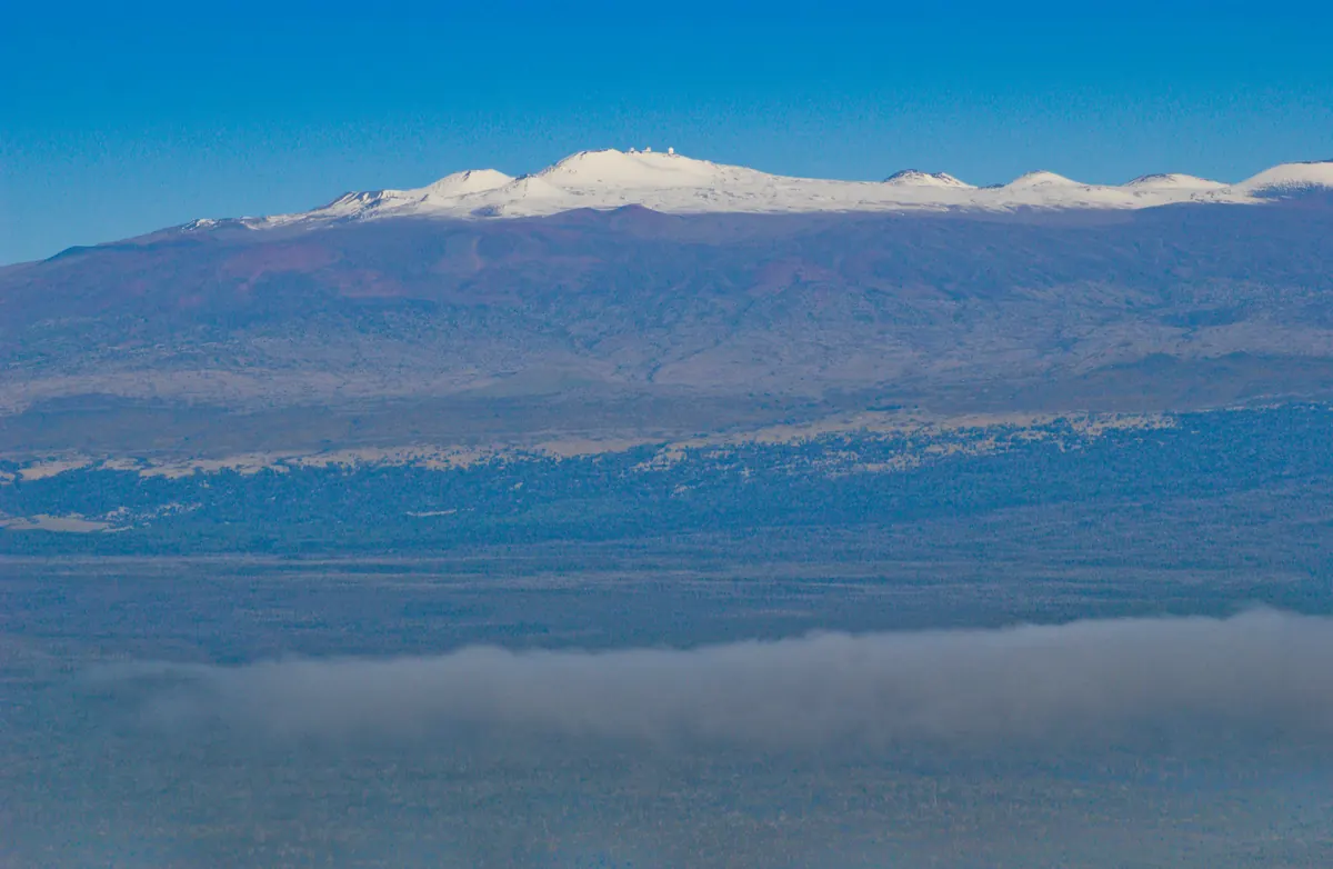 The broad summit of Maunakea capped with white snow above brown and reddish slopes, seen from the air with a band of cloud in the foreground.