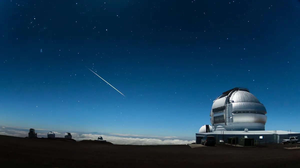 A bright meteor streaking across a starry sky above the Gemini North telescope dome on Maunakea, with the Pleiades low over other observatory domes to the left.