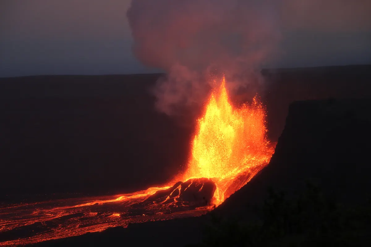Two tall lava fountains rising from vents on the floor of Halemaʻumaʻu crater, with the dark crater wall behind them.