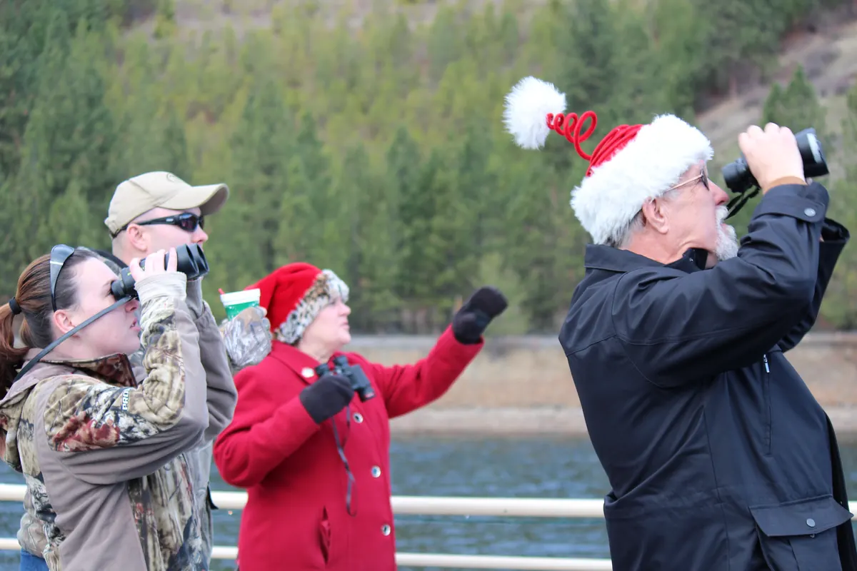 Four people in winter coats, two wearing red Santa hats, raise binoculars toward the sky beside a railing above a lake with forested hills behind.