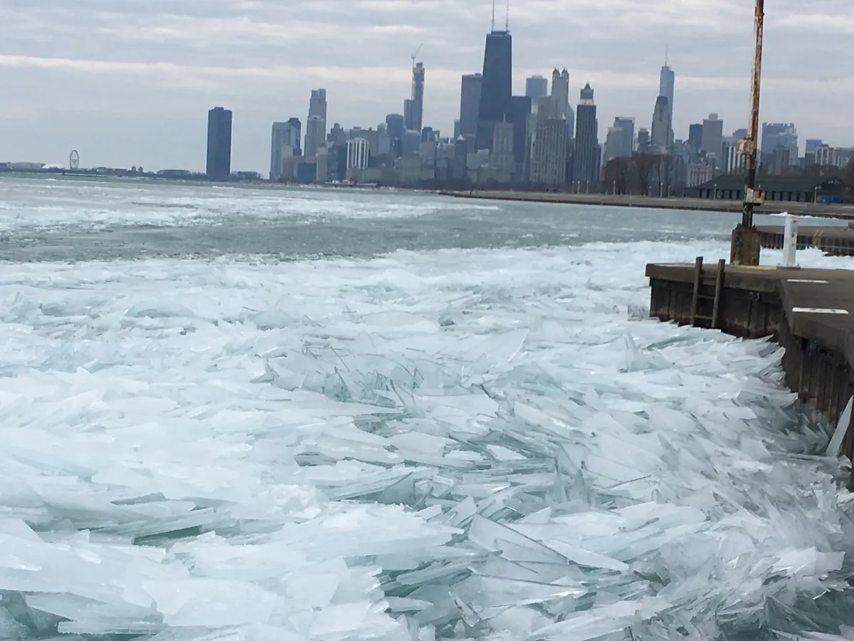 Broken plates of clear ice piled along a concrete breakwall on Lake Michigan, with the Chicago skyline in the distance under gray clouds