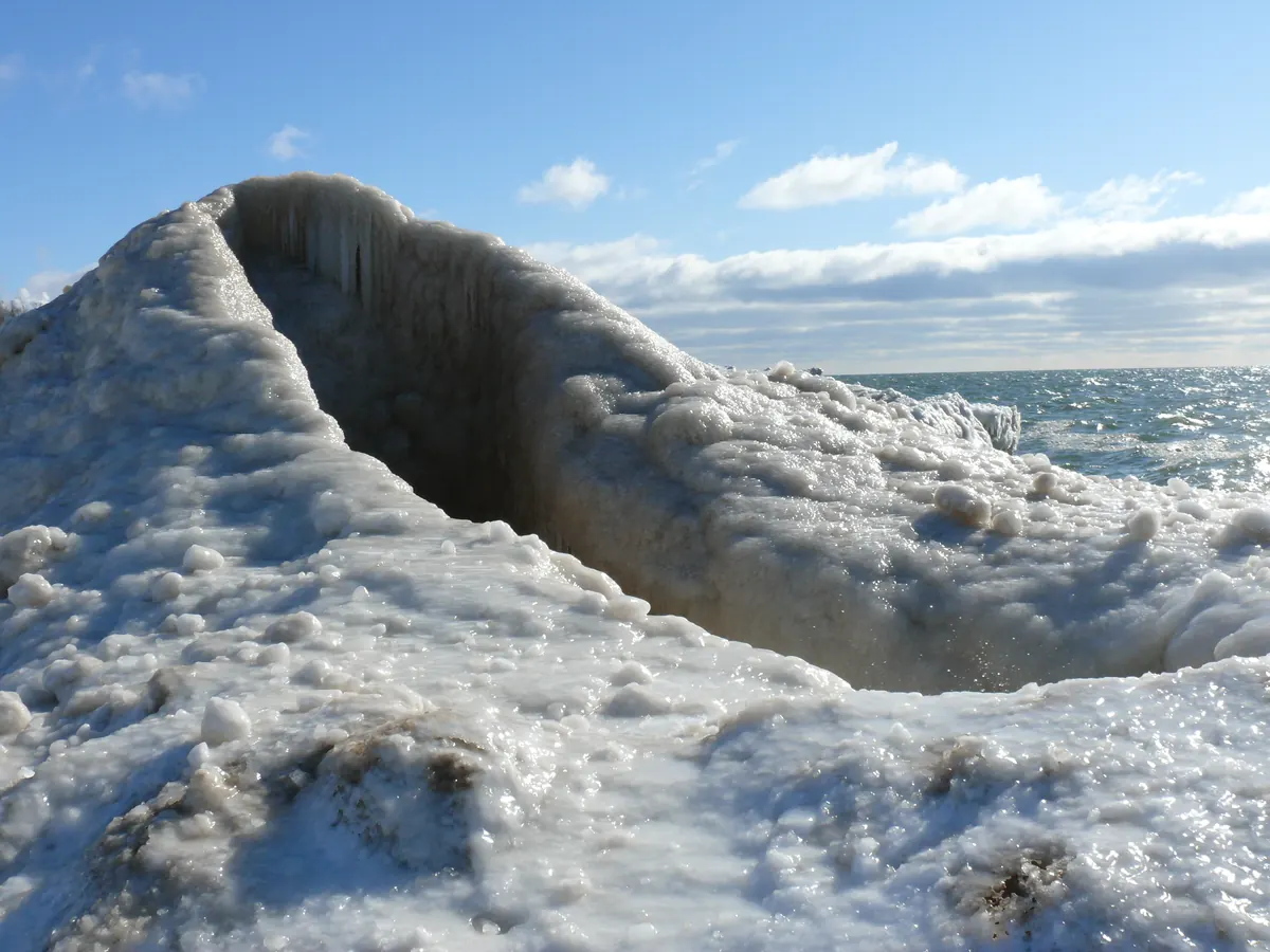 A tall, hollow mound of white ice on the Lake Michigan shore with open blue water and waves beyond it