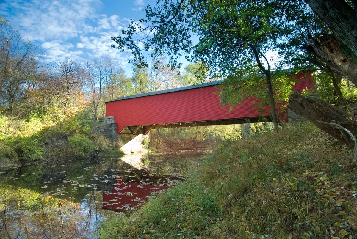 The red Ramp Creek Covered Bridge crossing a still creek, with autumn leaves on the water and turning trees along the banks.