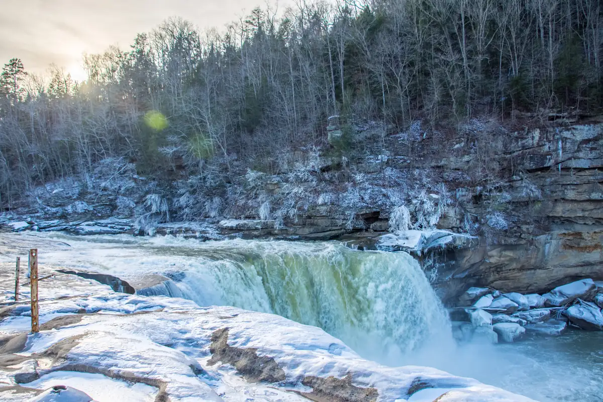 Cumberland Falls pours green-white water over a ledge between banks of snow and ice, with bare winter forest on the cliffs above.