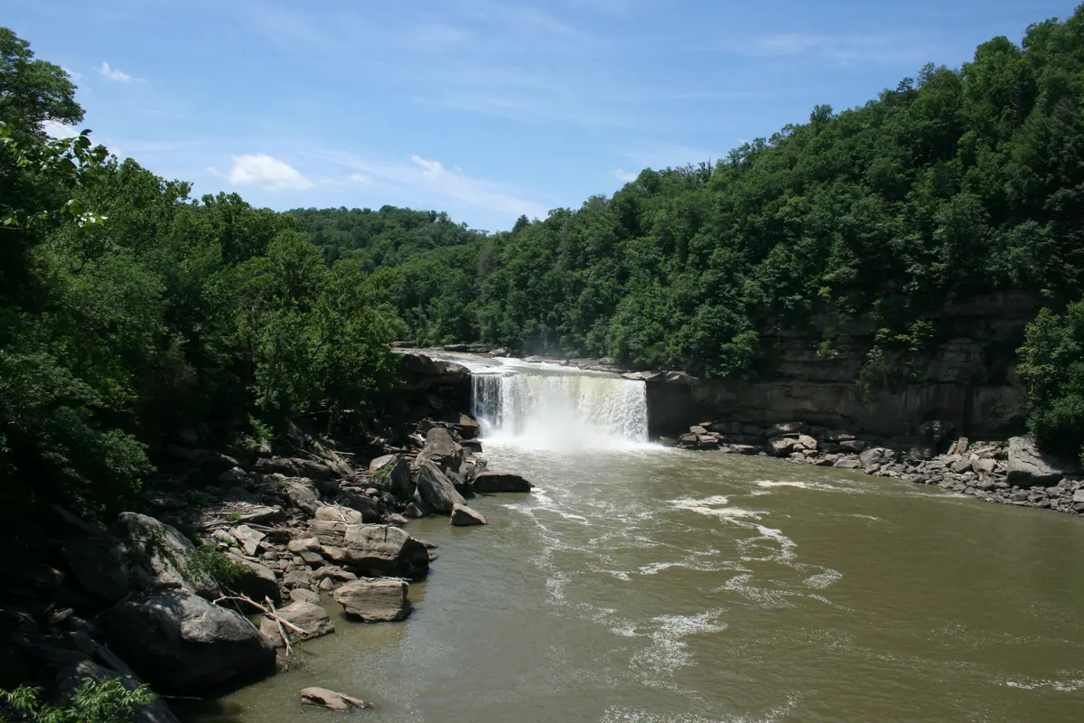 Cumberland Falls pours in a wide white curtain over a sandstone ledge into a broad river gorge lined with boulders and green forest.