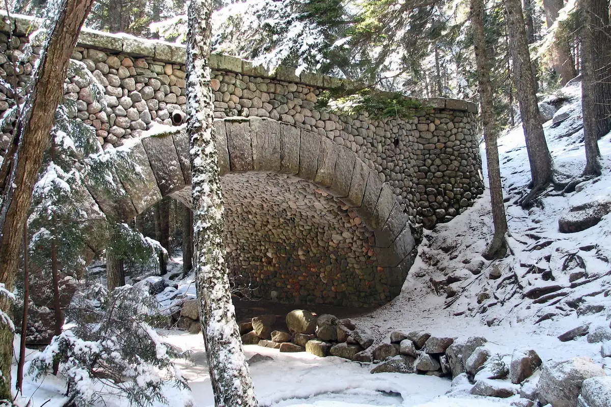 An arched bridge built of rounded fieldstones spanning a small stream in a snowy conifer forest, with snow on the boulders and branches.