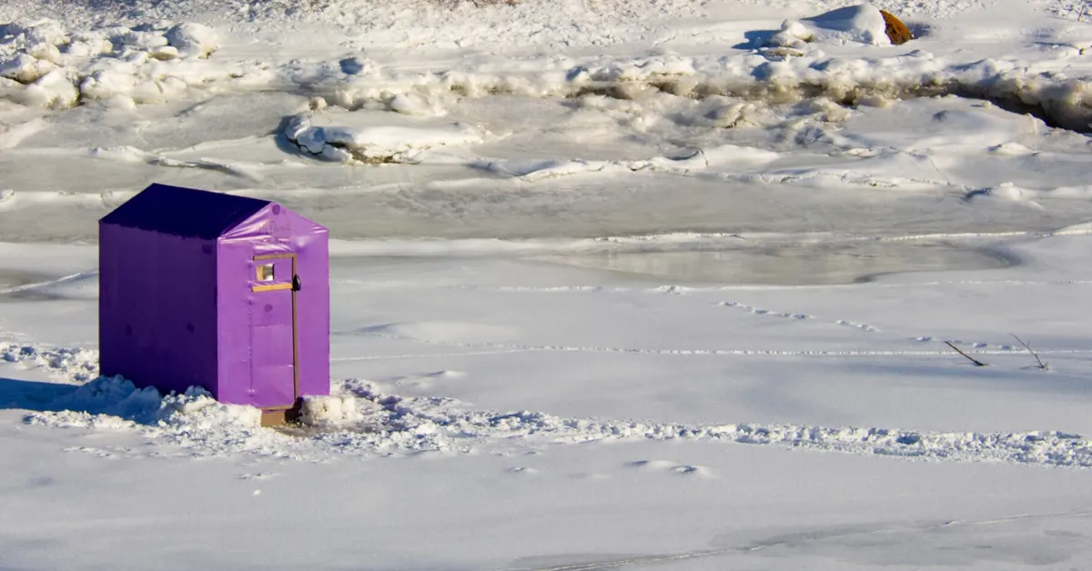 A small purple ice fishing shack standing alone on wide, snow-covered ice with jumbled ice behind it.