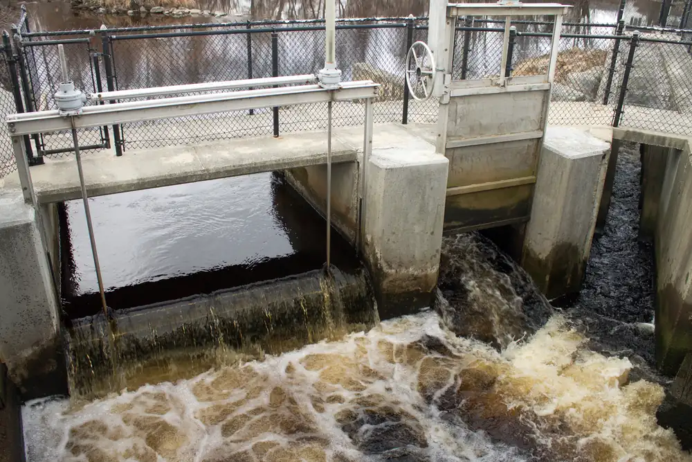 Dark, tannin-stained river water pouring over a concrete weir and through a small control gate beside a chain-link fence.