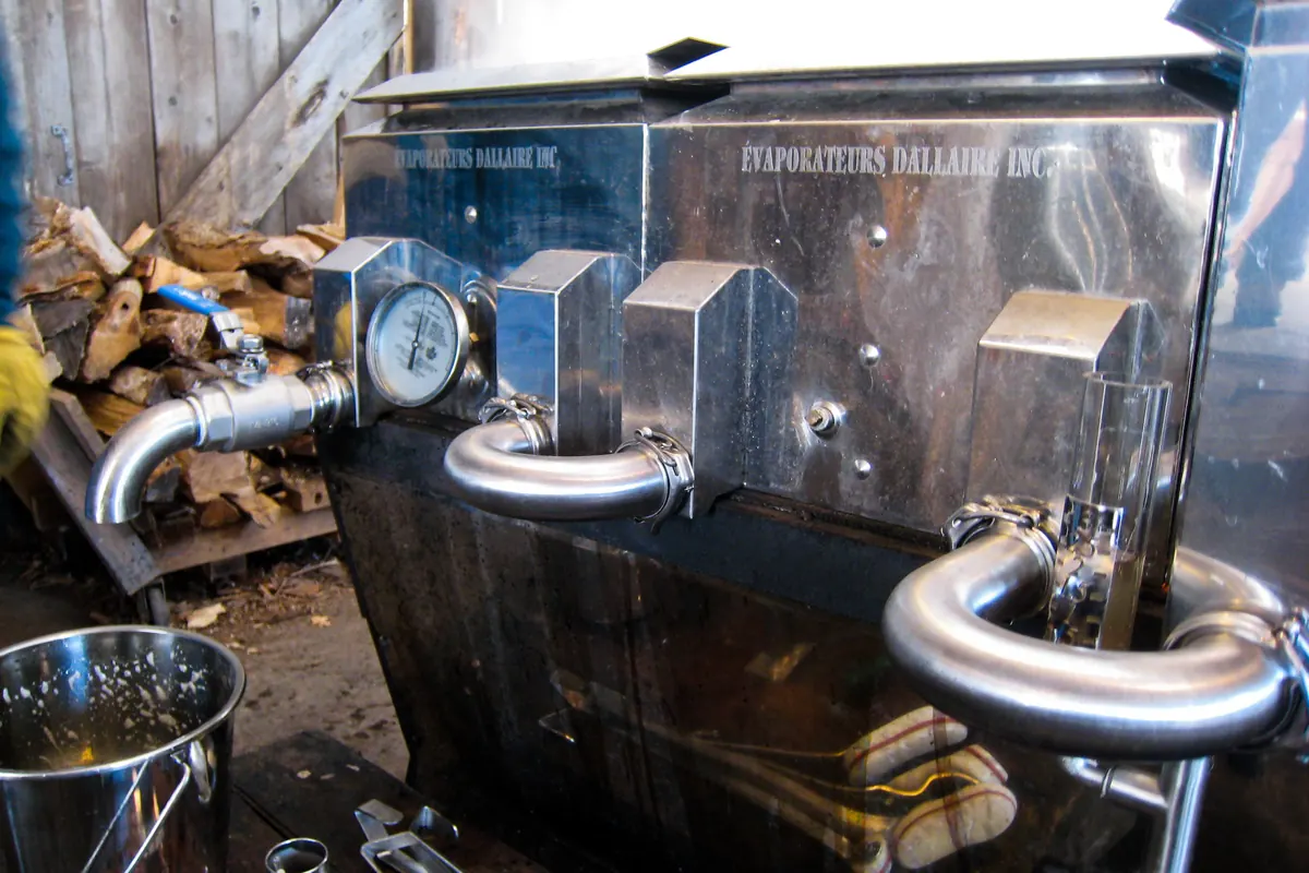 A stainless steel wood-fired maple syrup evaporator with a temperature gauge and curved pipes, a woodpile beside it in a plank sugarhouse.
