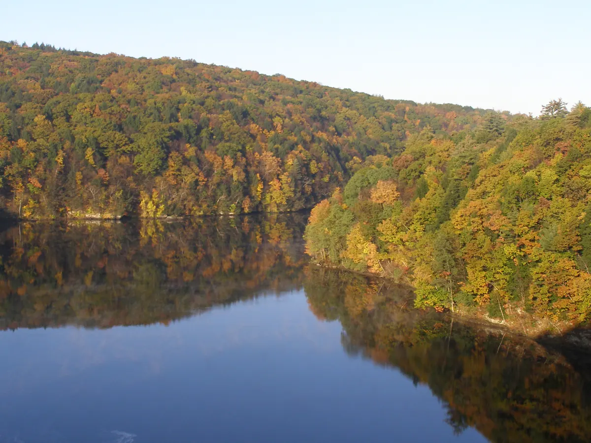 Wooded hills in orange, yellow and green fall color rising steeply from a calm river that reflects the trees and blue sky.