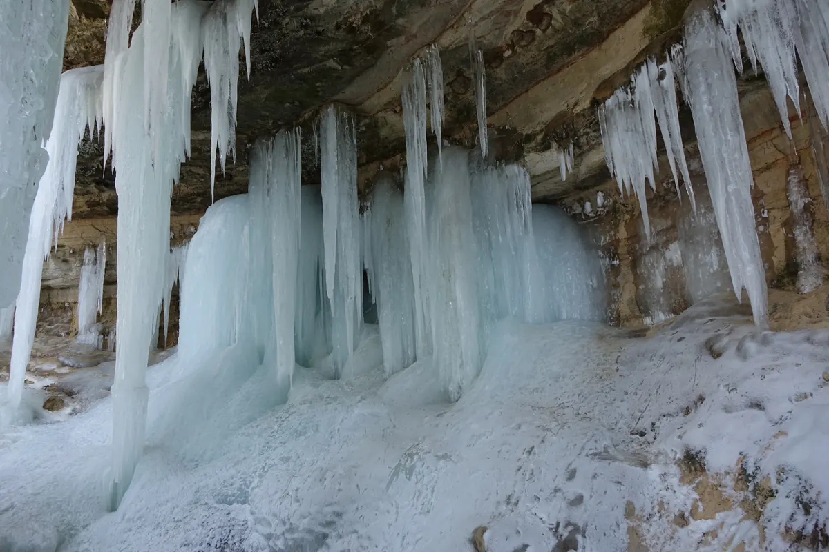 Long icicles and pale blue columns of ice hanging from a sandstone overhang above snowy ground