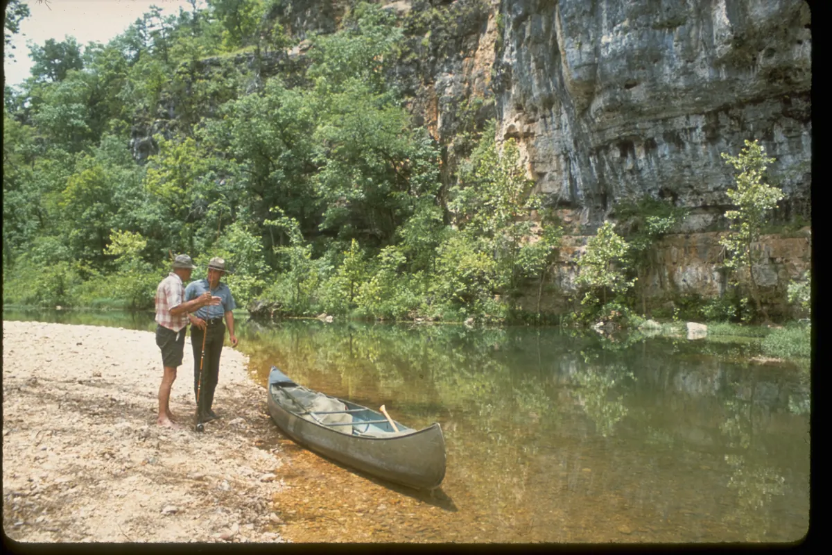 Two people standing on a gravel bar beside a beached canoe on a clear, shallow river beneath a tall limestone bluff