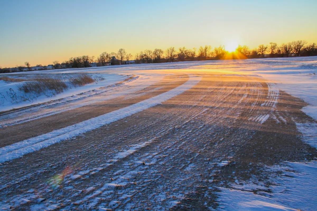 A low winter sun setting behind a line of bare trees at the end of a snow-covered gravel road in Nebraska