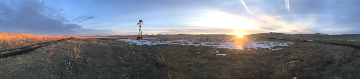 A wide view of tan Sandhills grassland with patches of snow and a windmill, the low sun setting under a streaky sky.