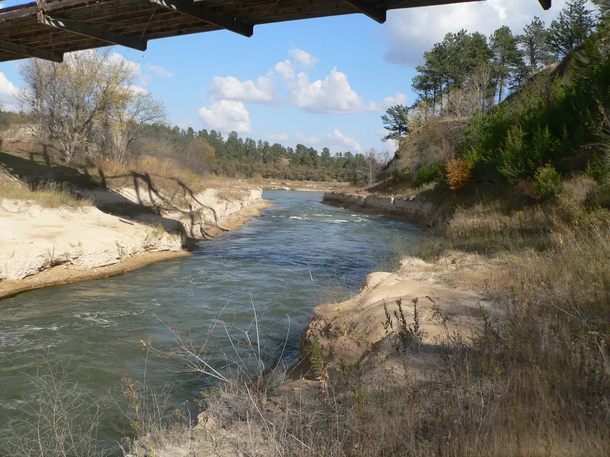 The shallow Niobrara River flowing east beneath the Norden Bridge between wooded bluffs and sandstone banks.