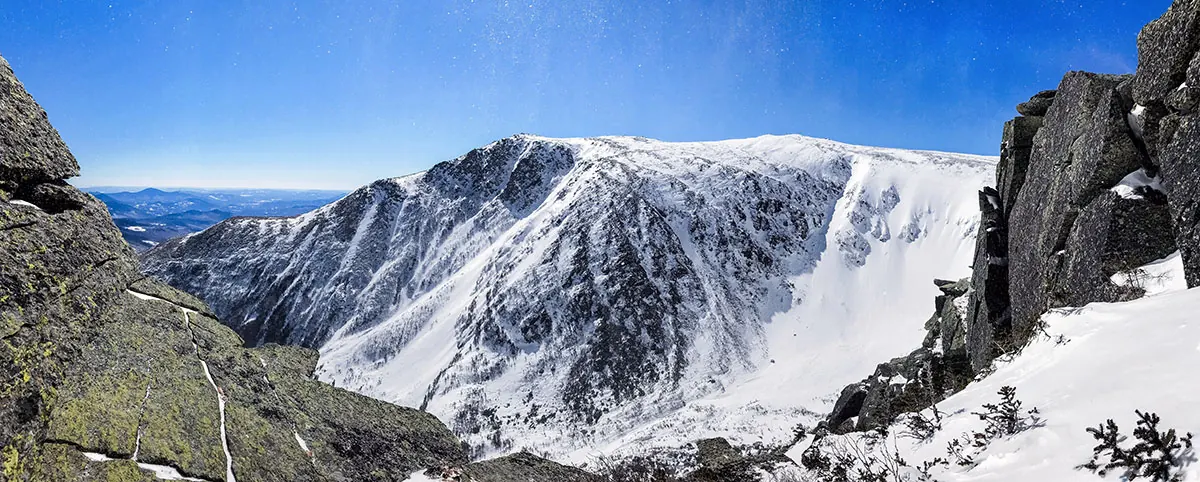 A snow-filled glacial bowl with dark rock cliffs on its walls, seen from rocky ledges under a deep blue sky, with forested valleys far below on the left.