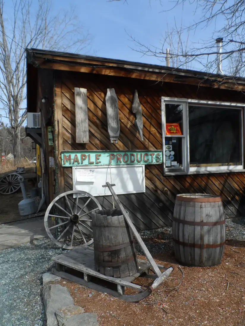 A weathered wooden sugar shack with a Maple Products sign, an old sap bucket on a sled, a wagon wheel and a barrel in front, under bare trees.