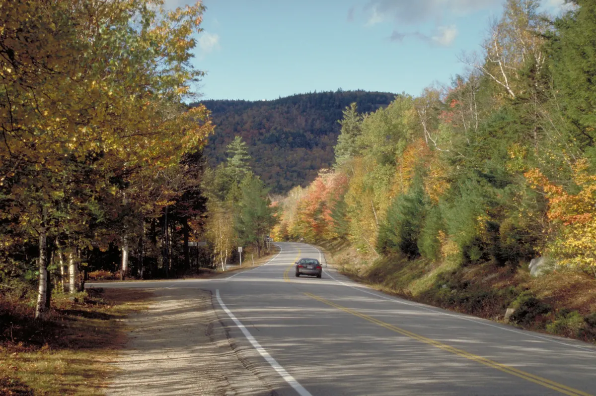A car travels a curving two-lane road lined with yellow birches, orange maples and dark conifers, with a wooded mountain rising ahead under a blue sky.