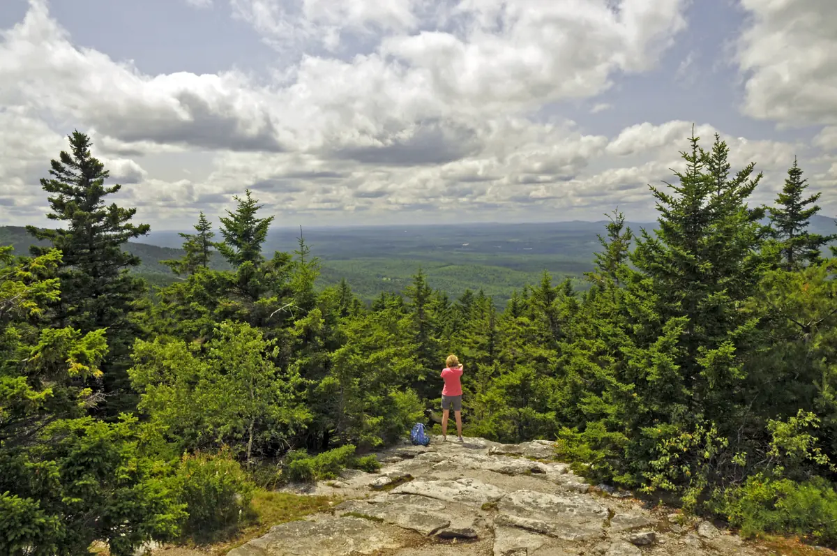 A hiker stands on a granite ledge among spruce trees, looking out over a wide forested valley under a sky full of cumulus clouds.