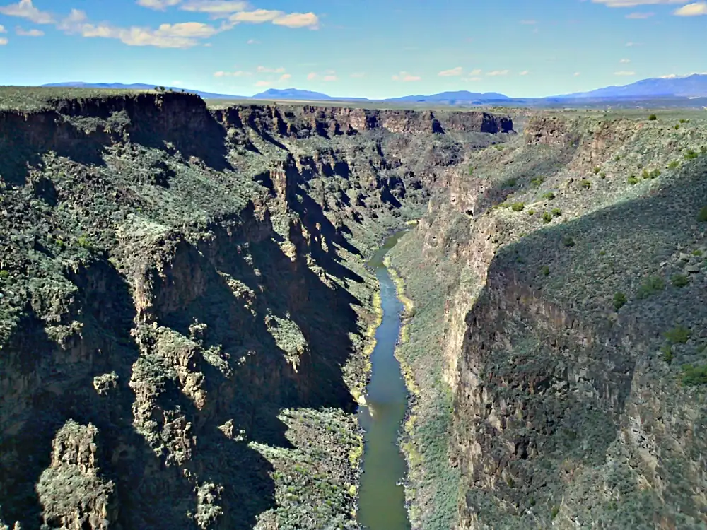 A narrow green river winding along the floor of a steep, shadowed gorge with sagebrush slopes, under blue sky with distant mountains