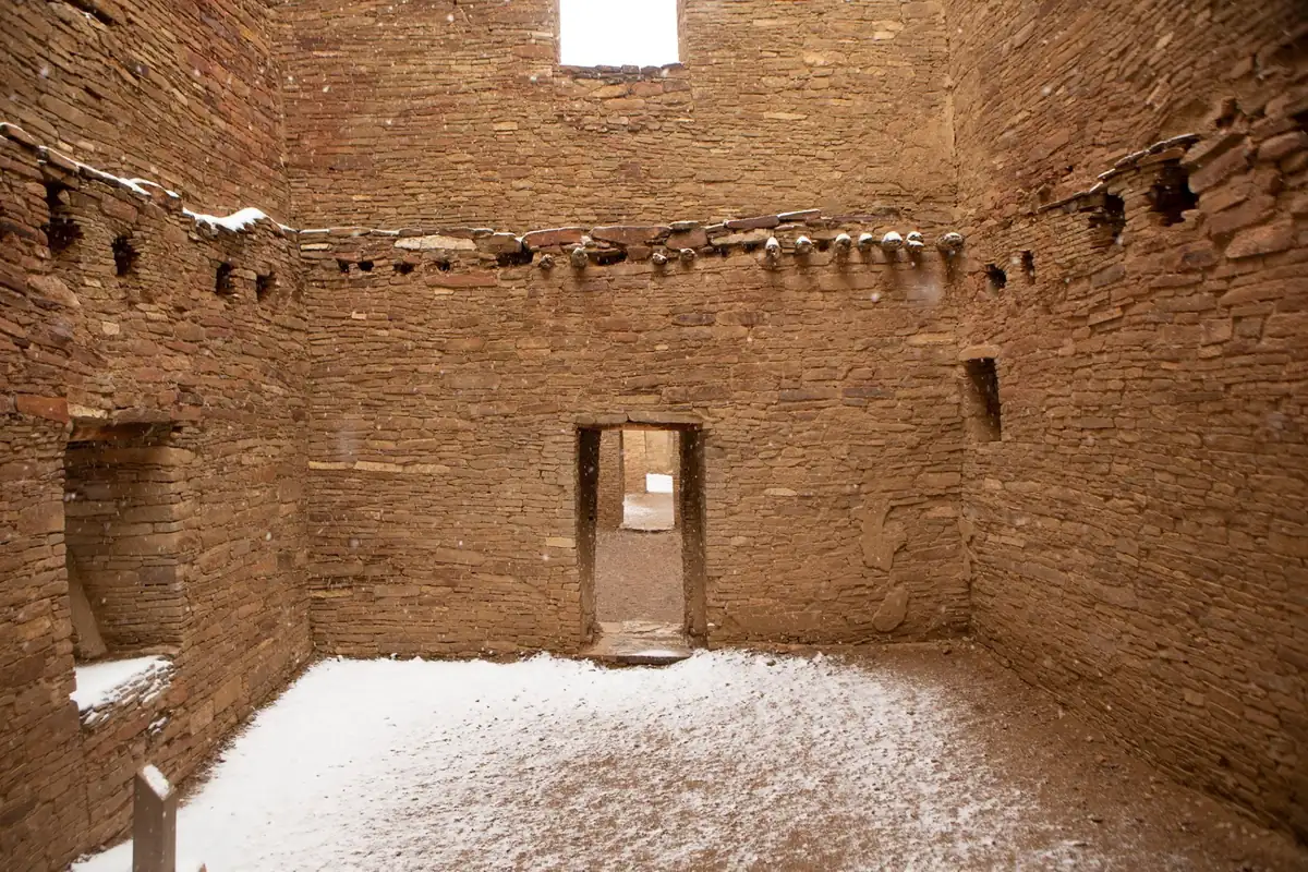 A roofless room of tall, finely laid sandstone masonry with a doorway in the far wall and a light dusting of snow on the floor.