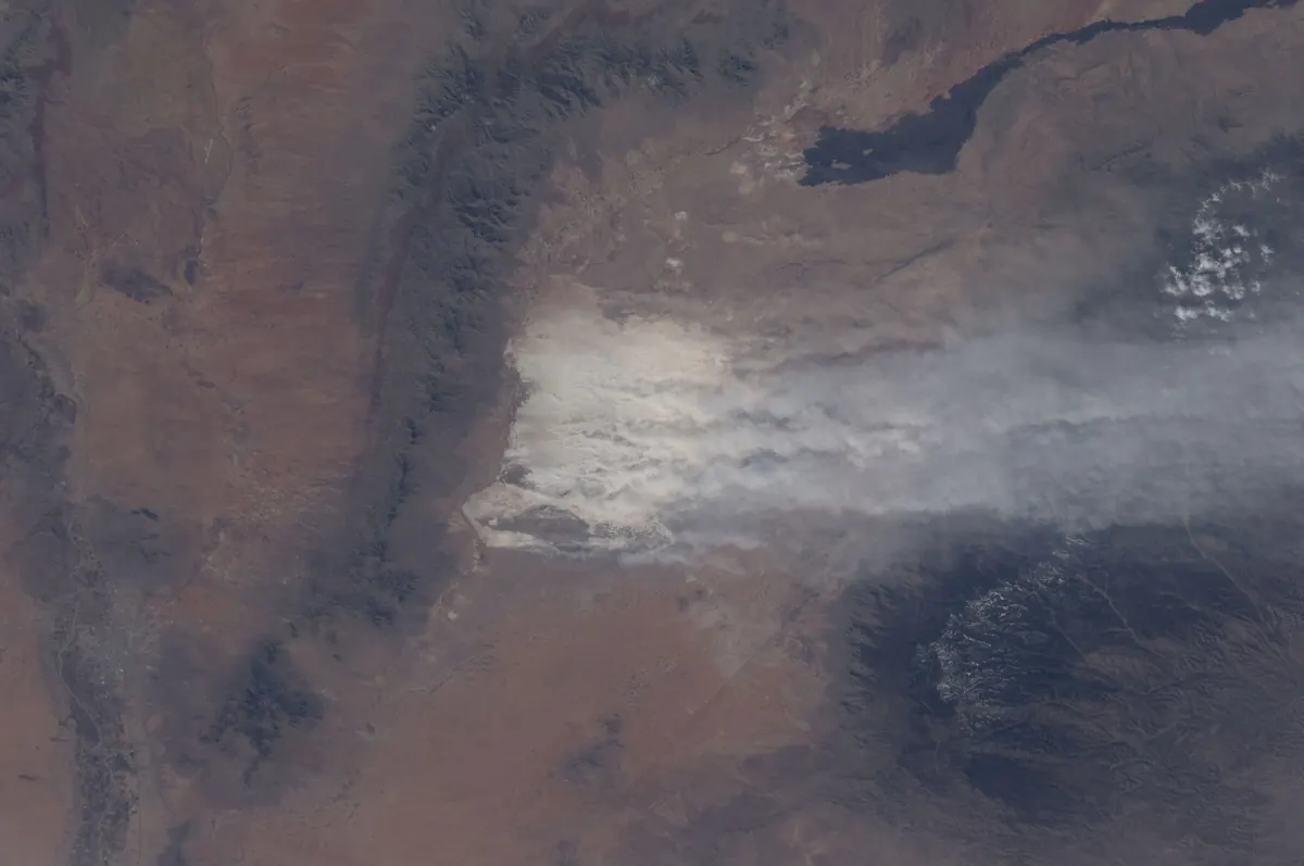 View from orbit of a pale plume of dust streaming east from a white dune field across brown desert and over dark, snow-dusted mountains