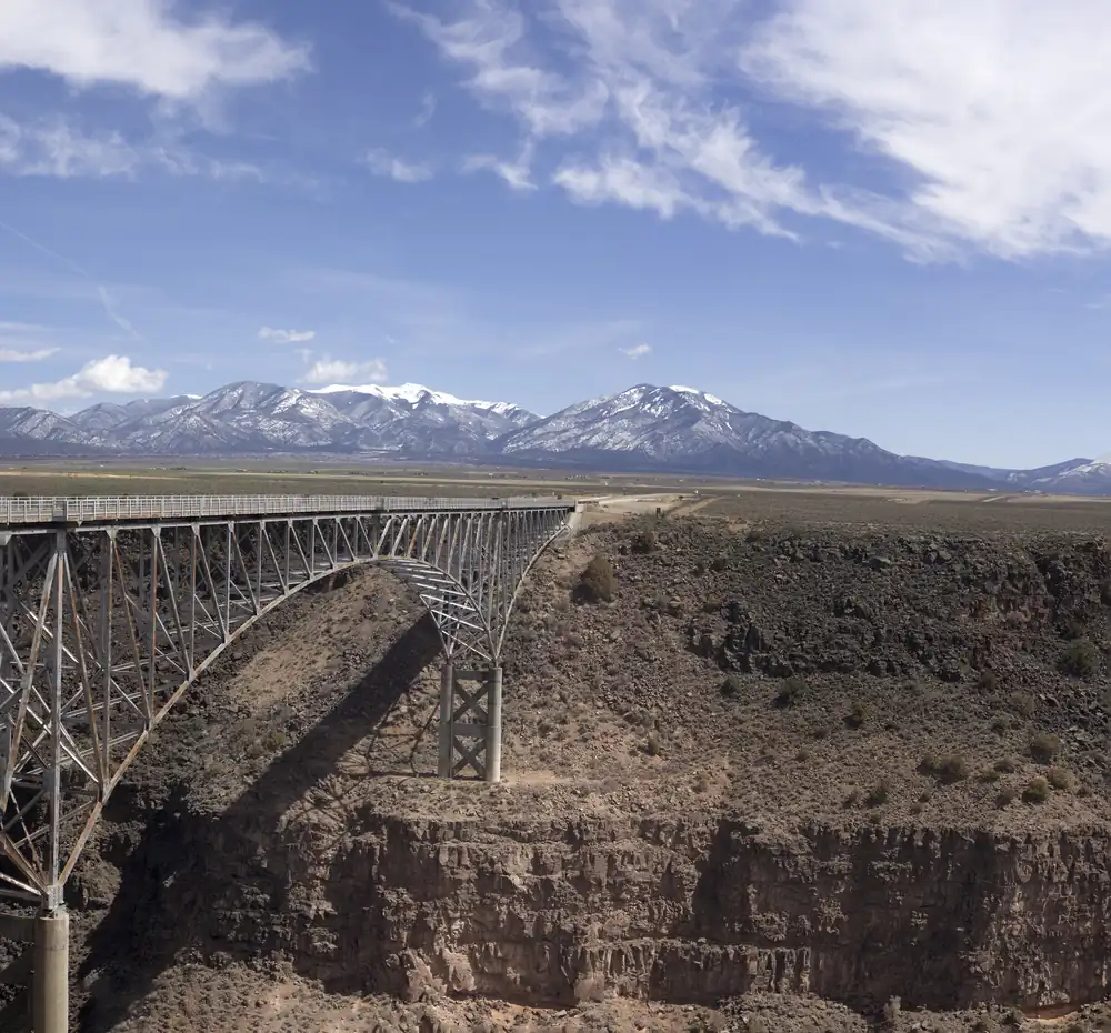 A steel arch bridge spanning a deep rock gorge, with snow-dusted mountains and blue sky beyond a flat sagebrush plain