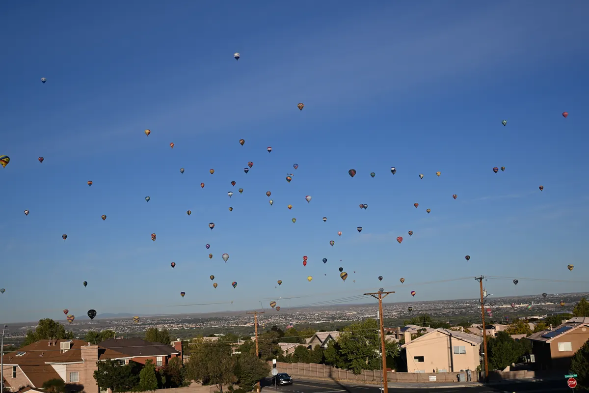 Dozens of hot air balloons scattered across a clear blue morning sky above rooftops and power lines on Albuquerque’s northeast side.