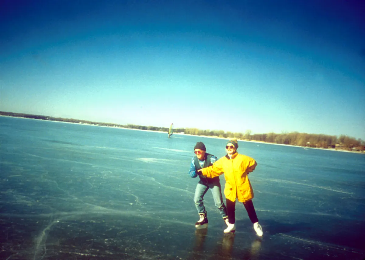 Two skaters in winter jackets glide across clear, cracked gray ice on a wide frozen bay under a deep blue sky, with a wooded shore behind