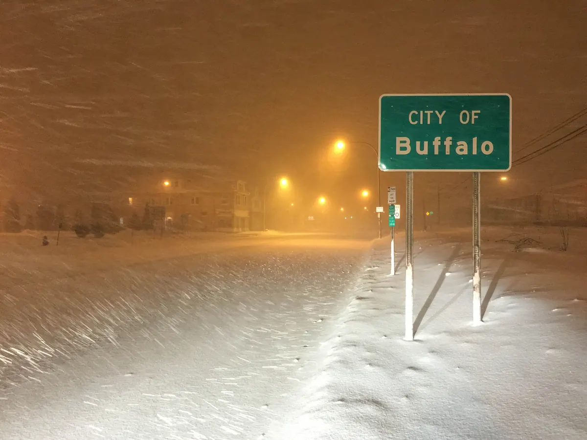 Heavy snow blowing across a drifted, empty road at night under orange streetlights, beside a green City of Buffalo sign.