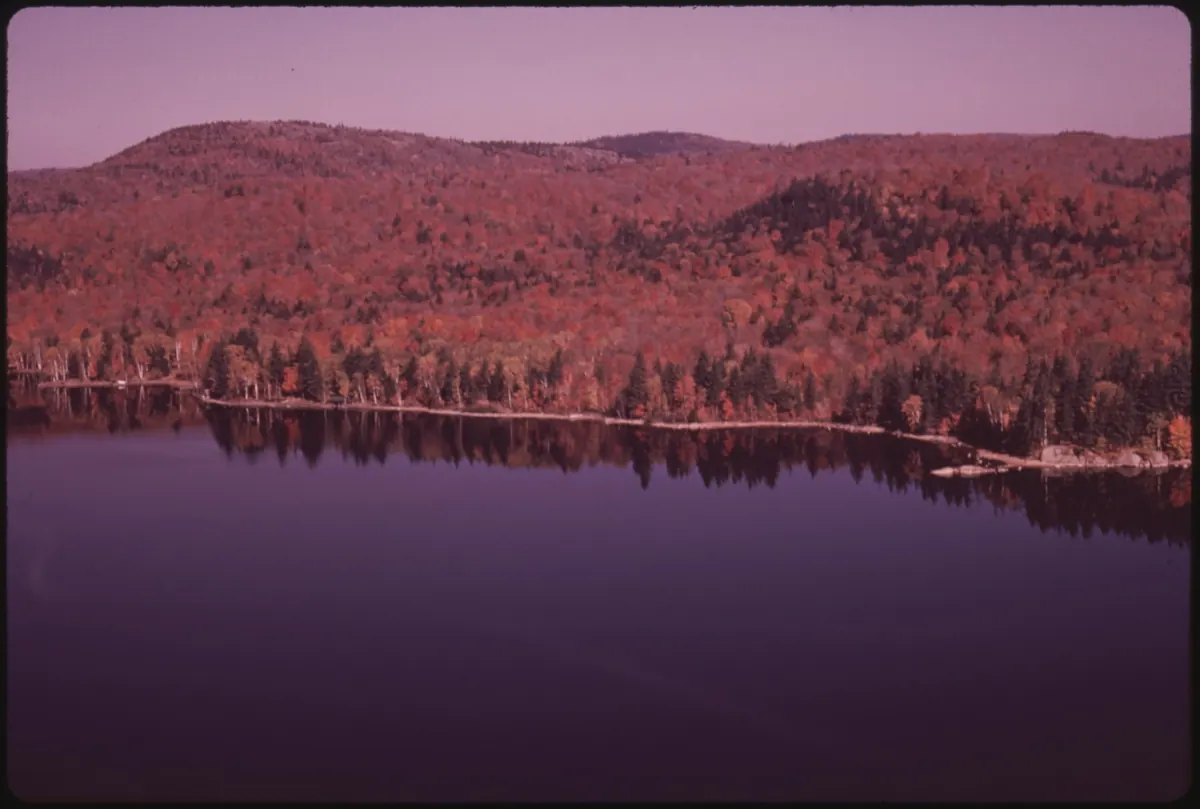 Hillsides covered in red and orange autumn hardwoods, with dark conifers along the shore, above the calm water of Seventh Lake in the Adirondacks.