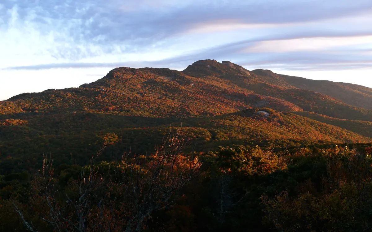 Low clouds spilling over the rocky peaks of Grandfather Mountain at sunrise, the slopes below covered in red, orange and gold fall foliage