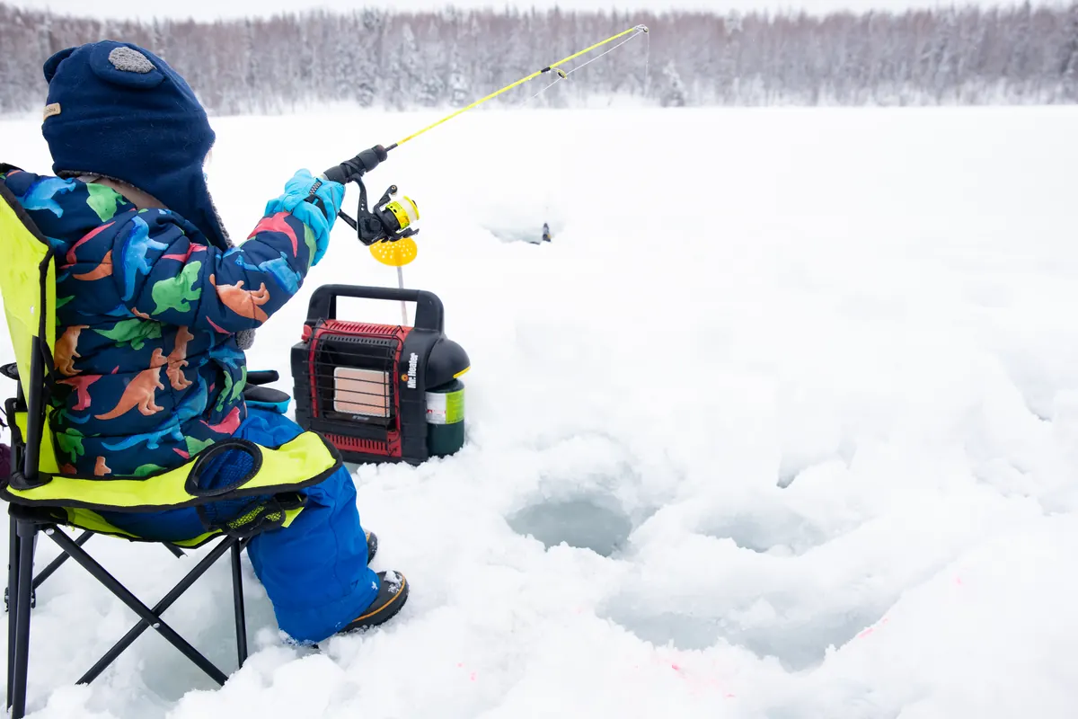 A small child in a snowsuit sitting in a camp chair beside a heater, holding a short fishing rod over a hole in snow-covered lake ice.