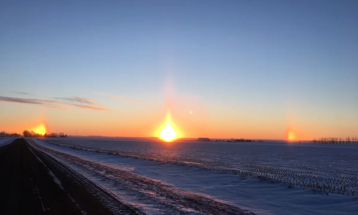 The rising sun over a snowy, flat field with a bright sundog on each side of it and a faint pillar of light above, under a clear orange and blue sky.