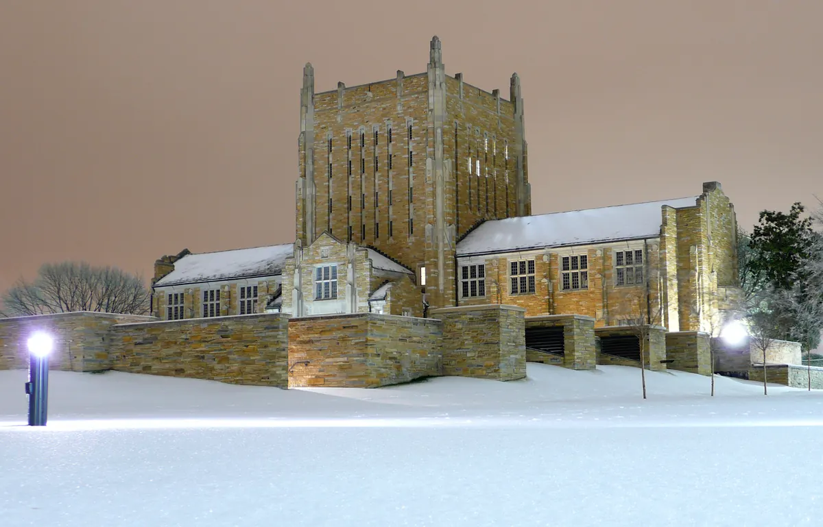 A tall stone library building at night with snow on its roofs and a smooth white blanket of snow and ice covering the lawn