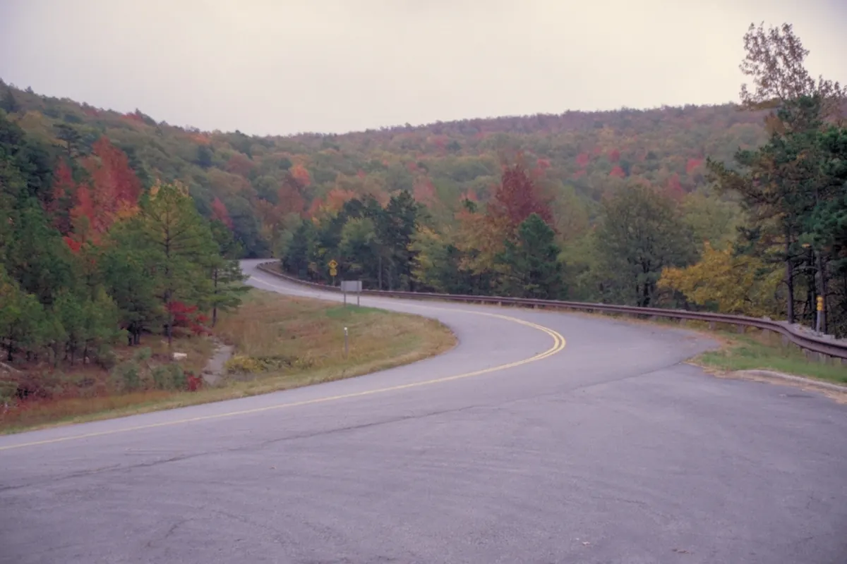 A curving two-lane road runs past forested ridges splashed with red, orange and yellow leaves among green pines.