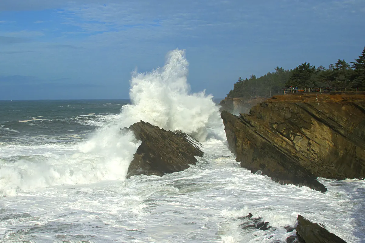 A huge white wave bursting over dark sandstone ledges below a forested bluff under a blue sky